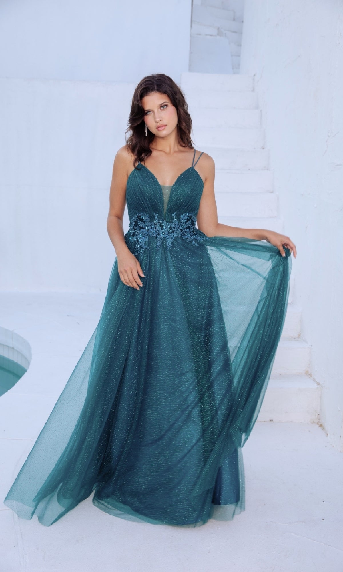 A woman in the Lace-Up Long Shimmer Prom Dress: Eureka 9797 stands on a white staircase, holding out part of her sleeveless teal A-line gown with a floral-embroidered waist.